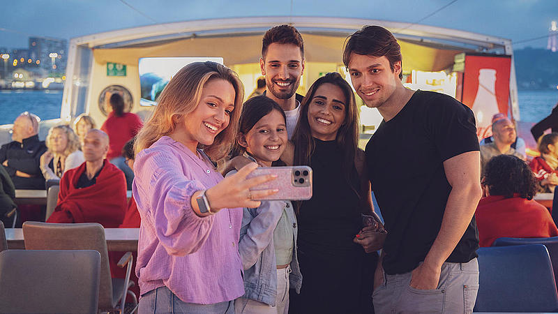 Un grupo de jóvenes se toman selfies en un crucero por el río al atardecer.