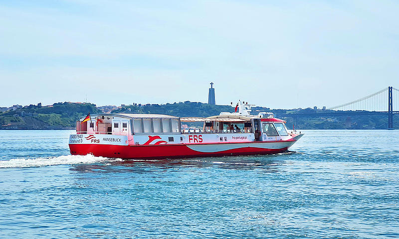Barco navegando por el río Tajo, con Cristo Rey y el Puente 25 de Abril al fondo.