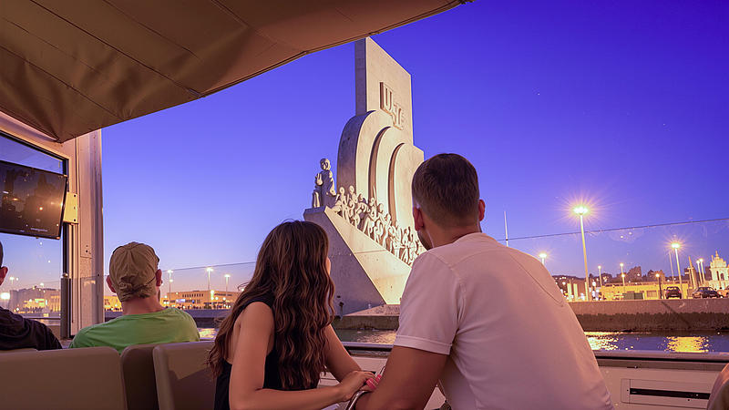 Paar auf einer Lissabon-Nachtfahrt blickt auf ein beleuchtetes Monument, mit Stadtlichtern im Hintergrund