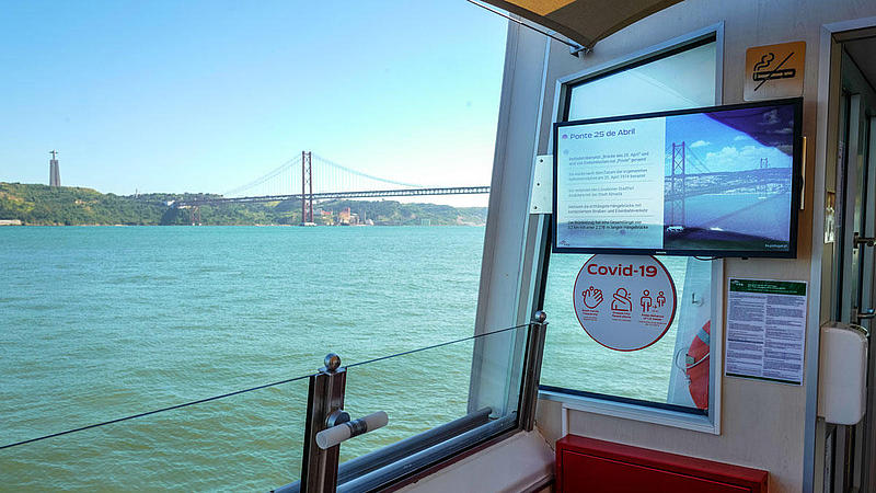 Bildschirm an Bord mit Infos über die Brücke des 25. April mit der realen Brücke im Hintergrund.